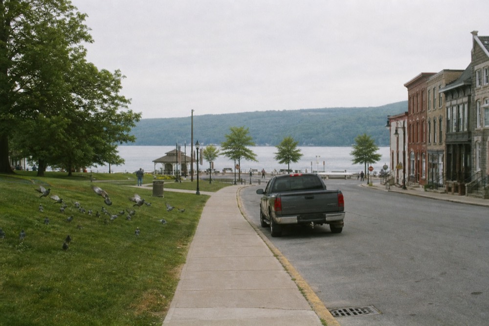 A village square at the head of a lake with historic storefronts, no text, no logos, no people